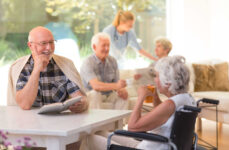 A group of seniors sitting socializing indoors engaged in various activities such as reading and conversing with each other.