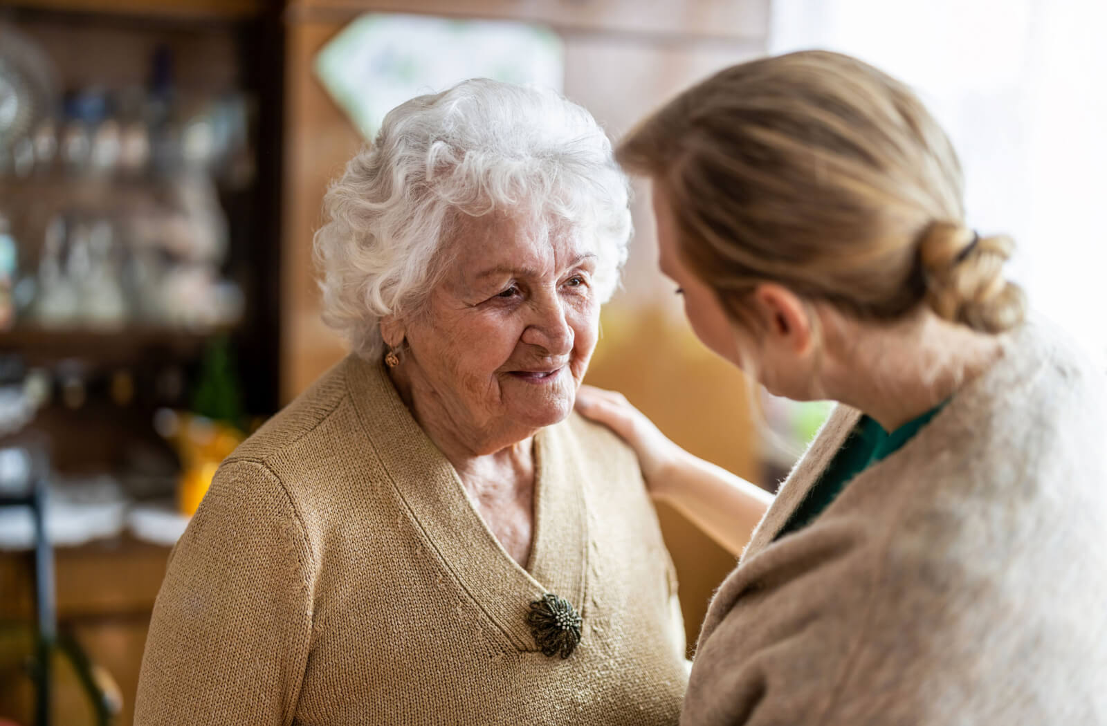 A young caregiver placing her hand on a senior woman's shoulder as she assists her in her day-to-day activities