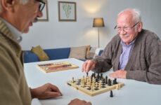 Two senior men sitting across from each other at a table playing a game of chess in a living room.