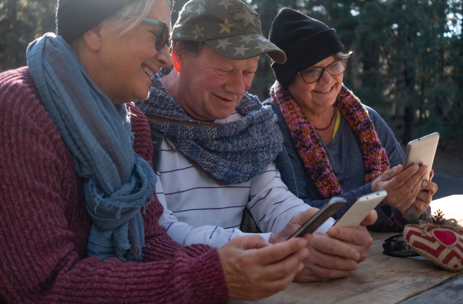 3 seniors sitting outdoors and wearing winter clothing while they use smartphones