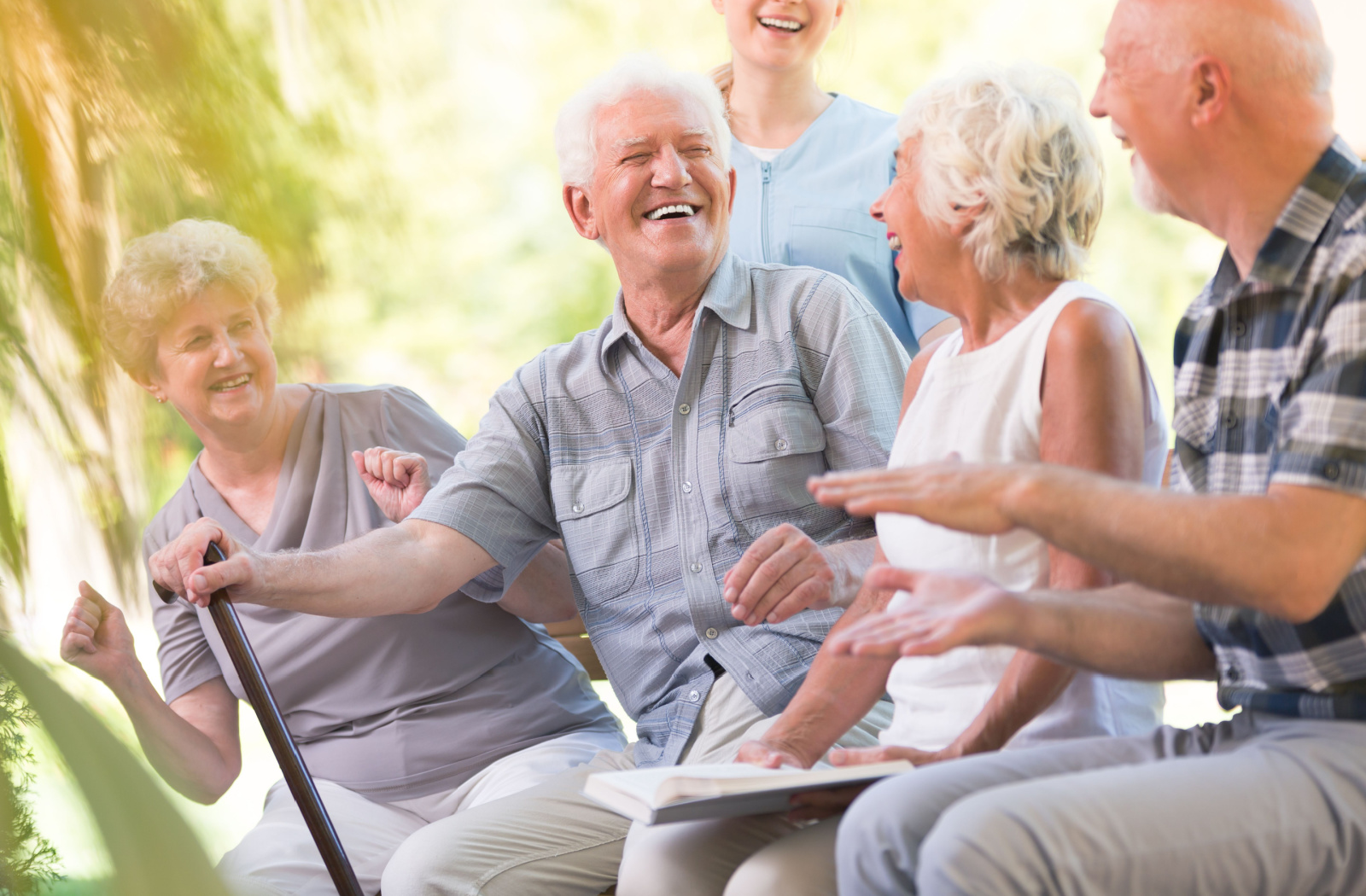 A senior man with a cane smiles and laughs while sitting outside with a nurse and other seniors.